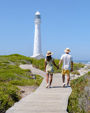 A couple holding hands walking a boardwalk path toward a lighthouse